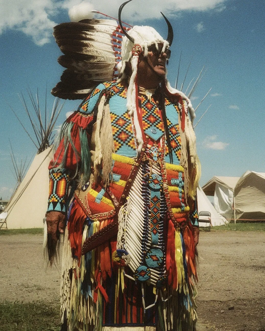 a man stands in traditional attire, adorned with horns, feathers and colorful fabrics and ribbons.