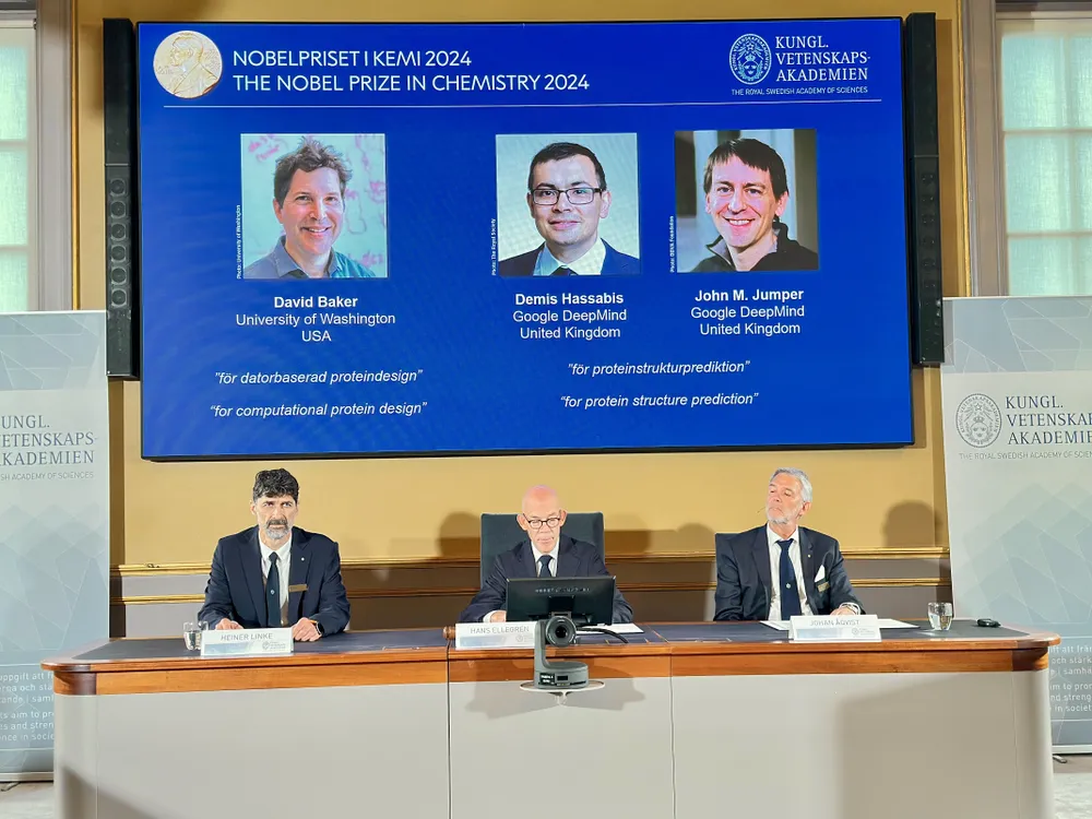 a slide showing the three Nobel winners is projected above three men sitting at a table