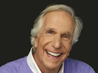 A headshot of Henry Winkler, shows a close-up the actor looking directly into the camera, his eyes twinkling above a broad, white smile.